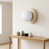 Wall sconce with a white globe shade and beige circular base mounted above a wooden console displaying small vases and a bowl.