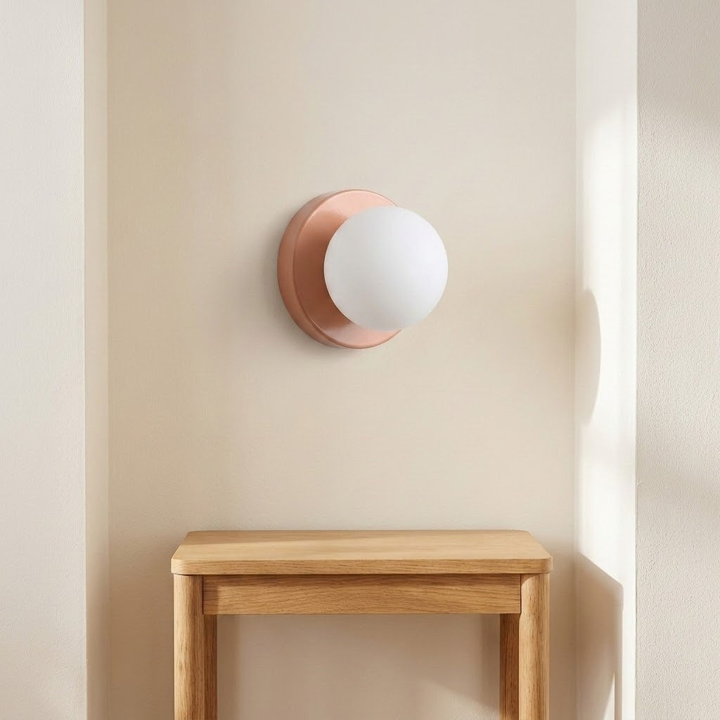 Wall sconce with a white globe shade and pink circular base mounted in a cream wall recess above a wooden table.
