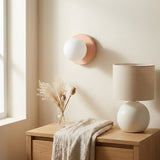 Wall sconce with a white globe shade and blush pink base above a wooden cabinet beside a window and table lamp.