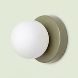 Wall sconce with a white plastic globe shade and muted green circular iron base against a pale green background.