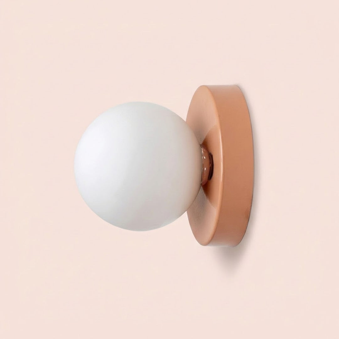 Wall sconce viewed from the side, with a white plastic globe shade and peach pink iron base against a pale pink background.