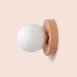 Wall sconce viewed from the side, with a white plastic globe shade and peach pink iron base against a pale pink background.