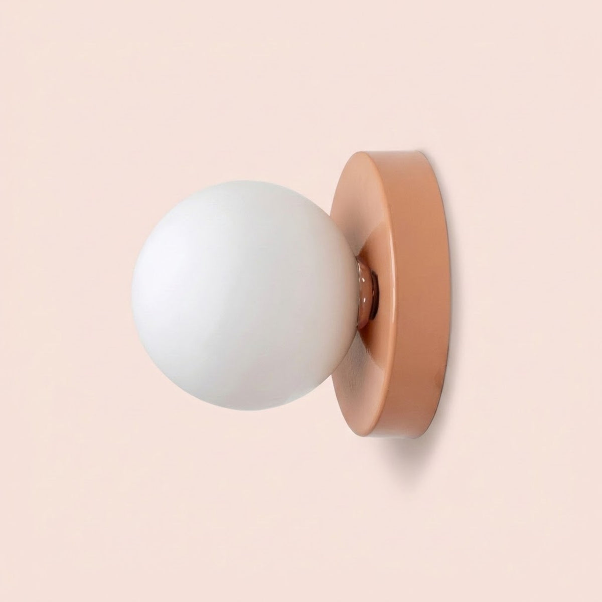 Wall sconce viewed from the side, with a white plastic globe shade and peach pink iron base against a pale pink background.