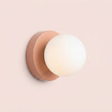Wall sconce with a softly glowing white plastic globe shade and peach pink circular iron base against a pale pink background.