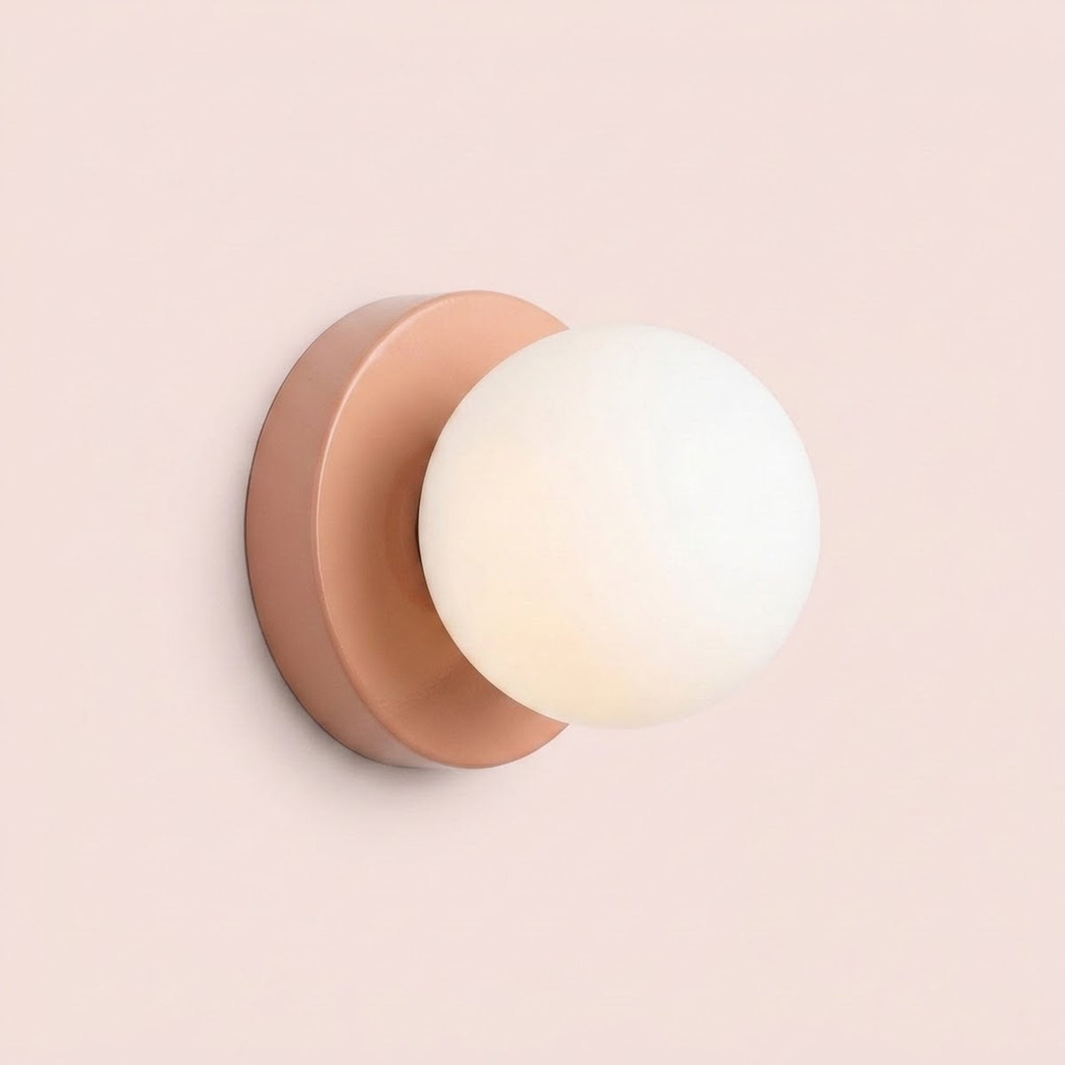 Wall sconce with a softly glowing white plastic globe shade and peach pink circular iron base against a pale pink background.