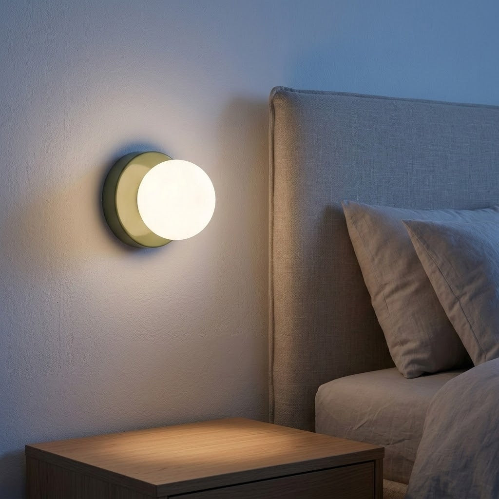 A wall sconce with a green base and glowing globe shade casts warm light beside an upholstered headboard and wooden bedside table.