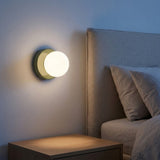 A wall sconce with a green base and glowing globe shade casts warm light beside an upholstered headboard and wooden bedside table.