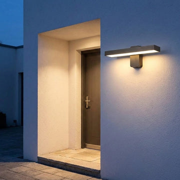 Dark rectangular wall sconce casts warm white light across a textured exterior wall and recessed doorway at dusk.
