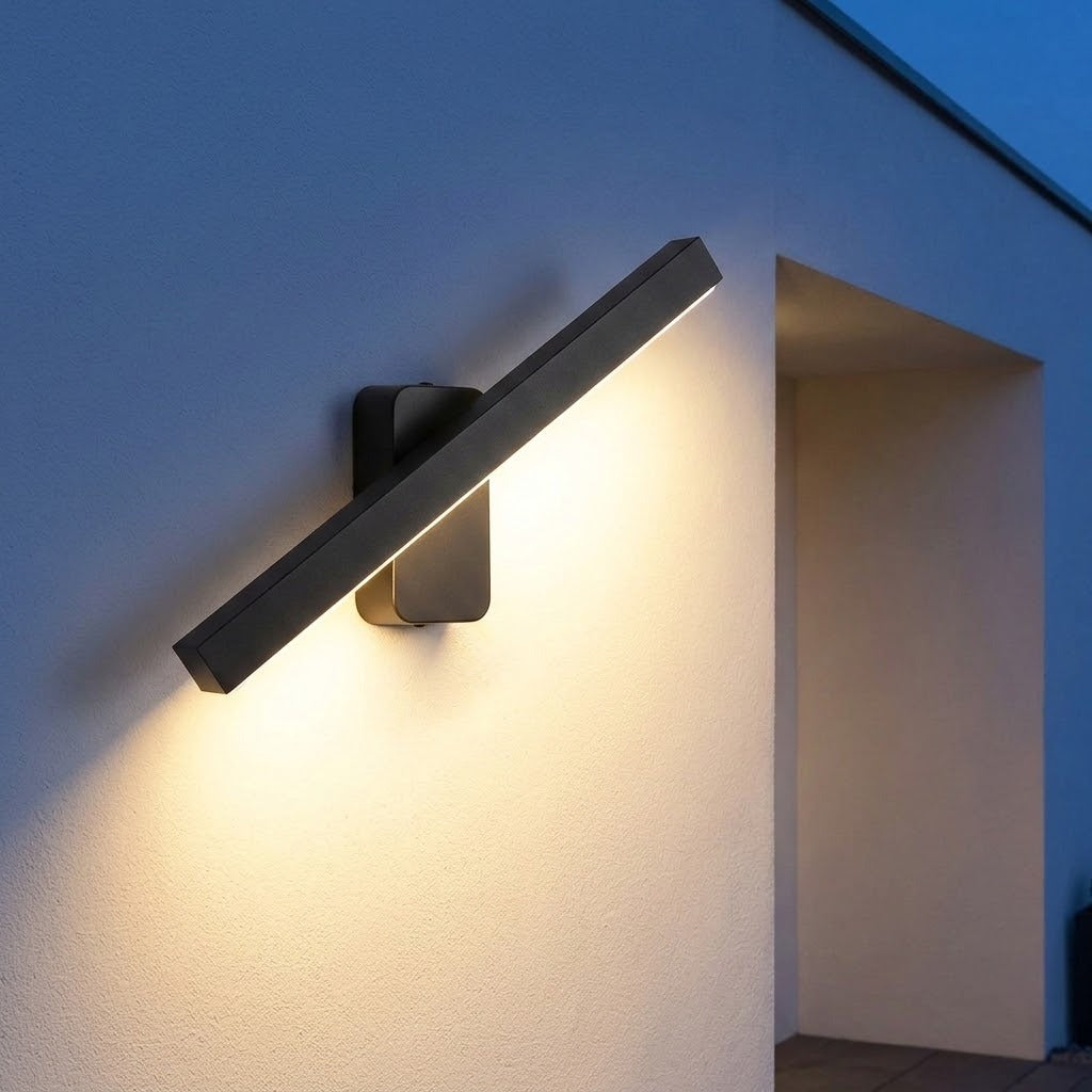 Modern rectangular wall sconce light fixture mounted on a textured wall, casting a warm yellow glow downward and outward. The fixture has a matte black finish and is shown in a real-world architectural setting at dusk.