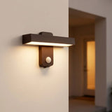 A modern rectangular wall sconce light fixture in a dark brown finish with an integrated motion sensor, mounted on a beige wall and casting a warm glow, with a softly blurred hallway in the background.
