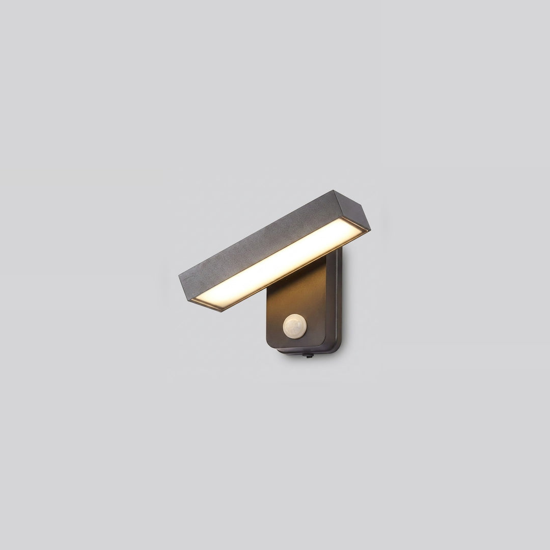 Dark gray wall sconce with an angled rectangular light bar glowing warm white and a round motion sensor against a pale gray background.