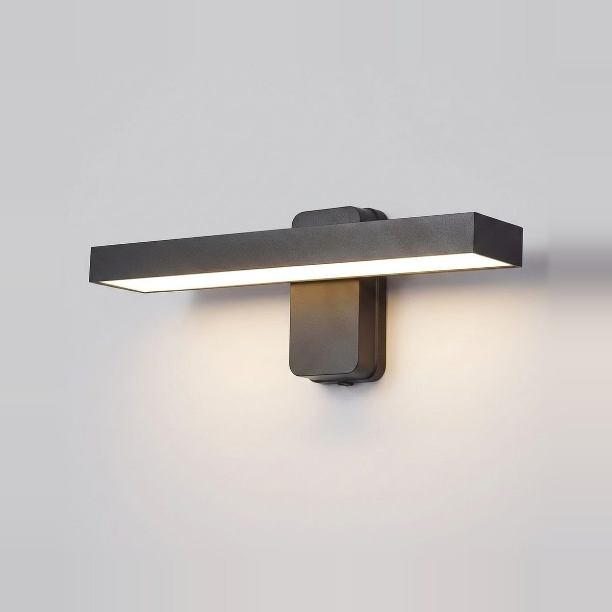 A modern horizontal wall sconce light fixture in a matte dark gray finish, mounted on a plain light gray wall and casting a warm glow downward.
