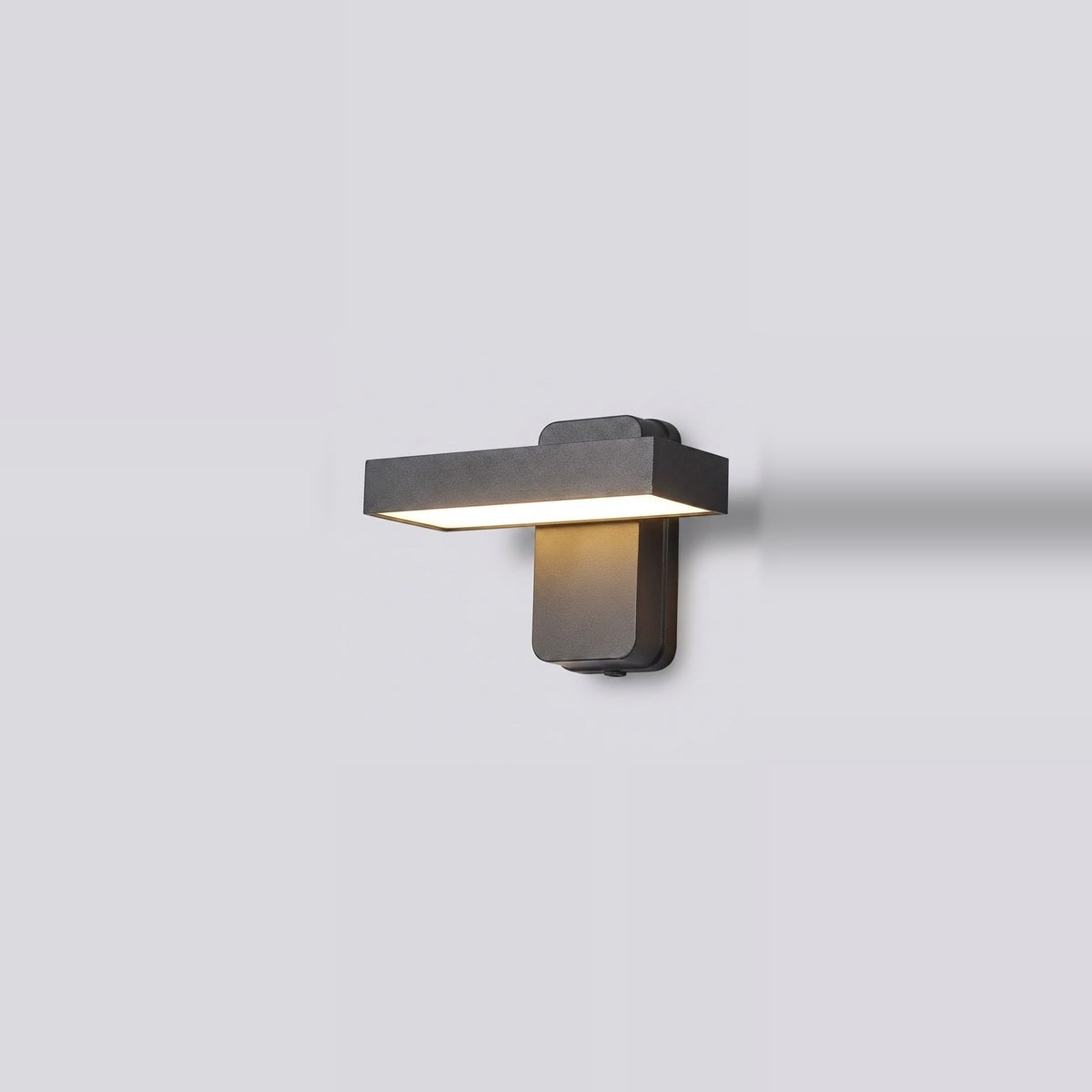 A modern rectangular wall sconce light fixture in a dark gray finish, mounted on a light gray wall and casting a warm yellow light downward.