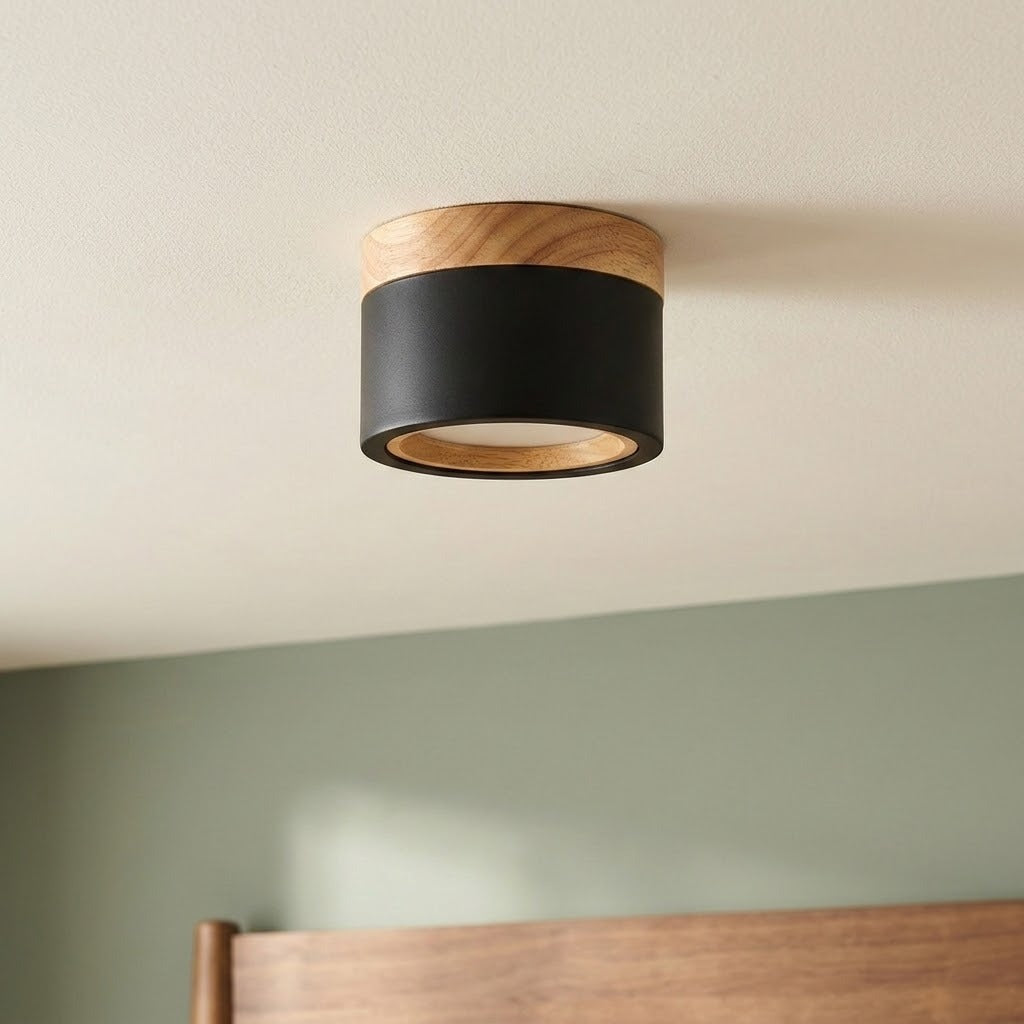 A black cylindrical LED downlight with wood trim mounts on a white ceiling above a green wall and wooden headboard.