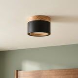 A black cylindrical LED downlight with wood trim mounts on a white ceiling above a green wall and wooden headboard.