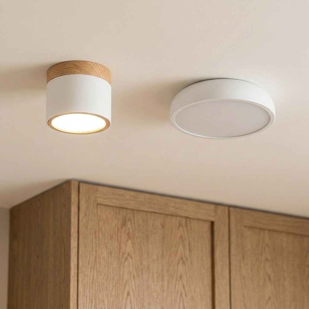 A lit white LED downlight with wood trim sits beside a flat round ceiling light above wooden kitchen cabinets.