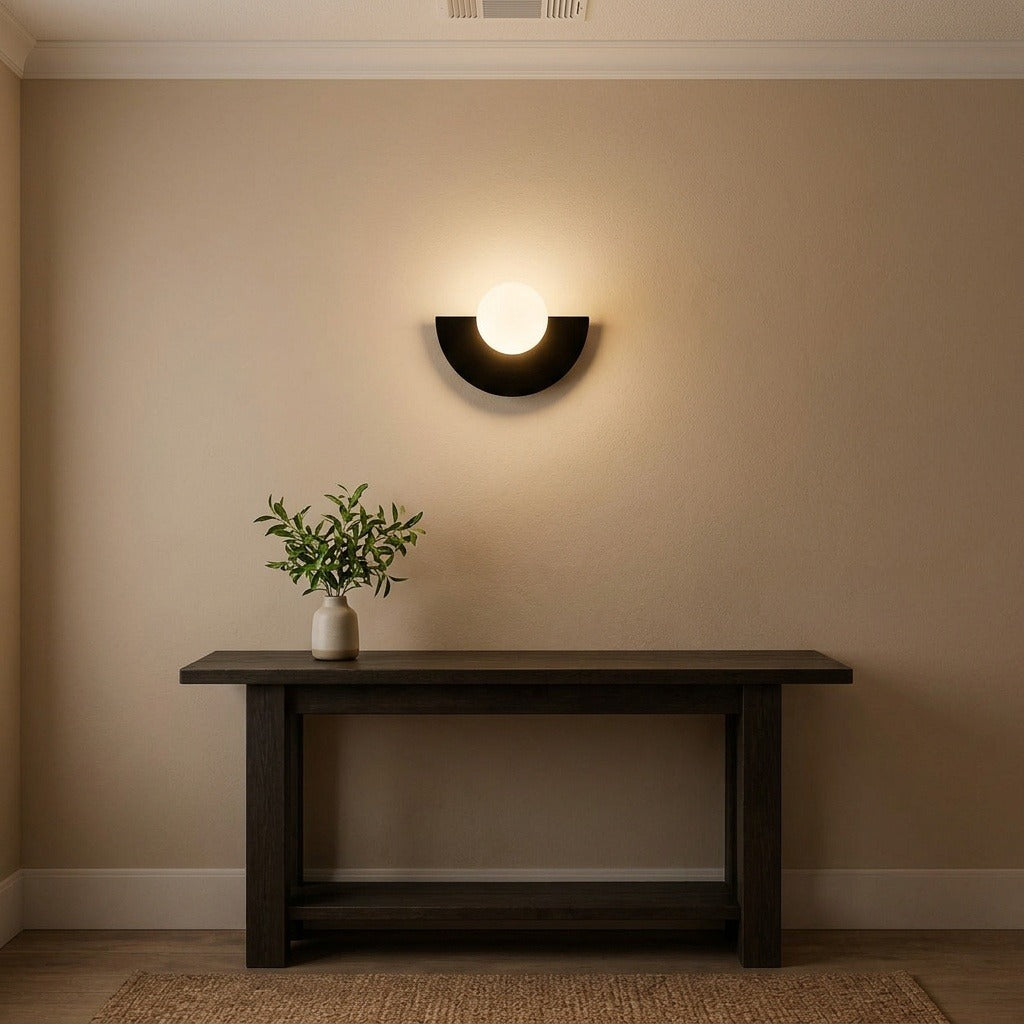 A modern black semi-circular wall sconce with a glowing white globe light, mounted on a beige wall above a dark wood console table with a small potted plant.
