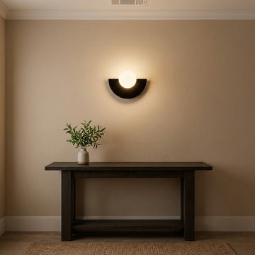 A modern black semi-circular wall sconce with a glowing white globe light, mounted on a beige wall above a dark wood console table with a small potted plant.