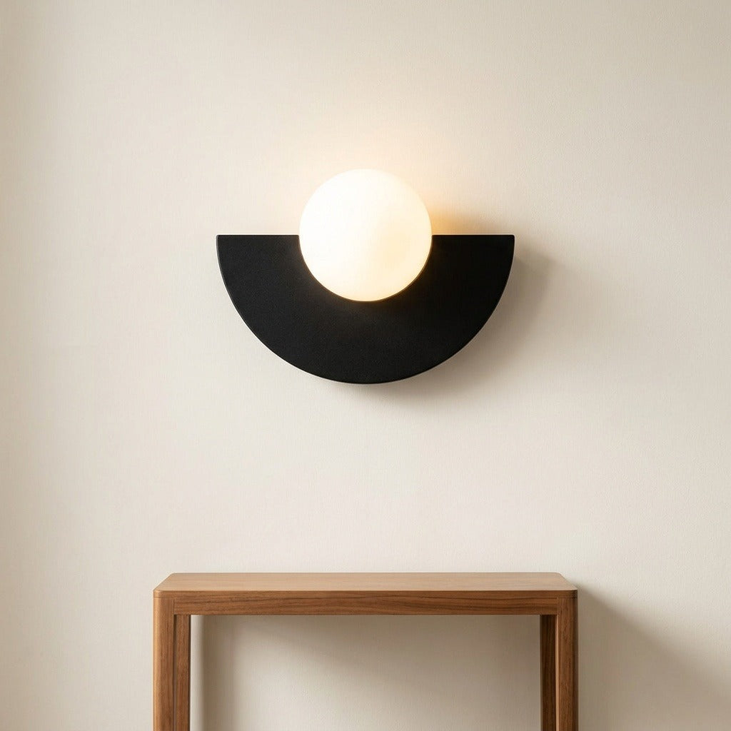A modern semi-circular wall sconce with a glowing white globe bulb, mounted on a light beige wall above a wooden console table.