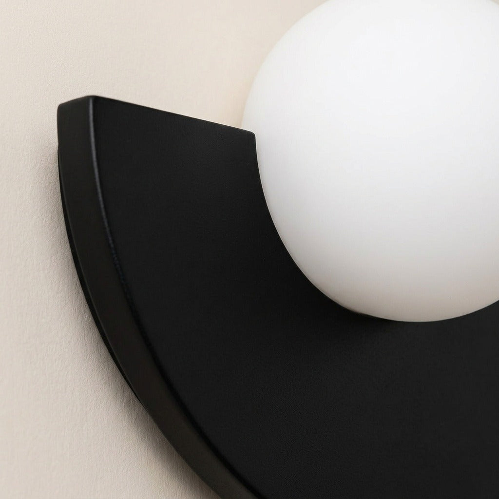 Close-up of a modern wall sconce with a matte black semi-circular frame and a large white spherical shade, mounted against a light beige wall.