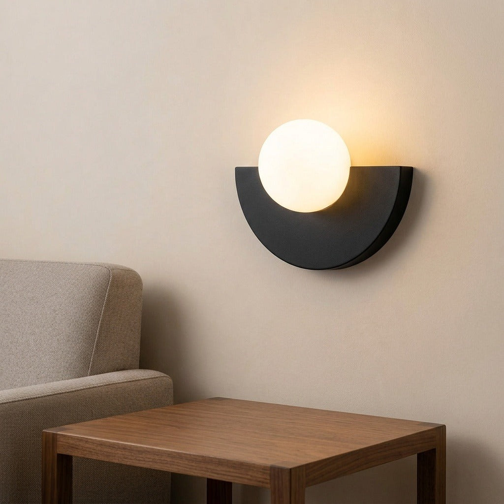 A modern semi-circular wall sconce with a glowing frosted white globe and a matte black half-moon frame, mounted on a beige wall above a wooden side table next to a gray sofa.
