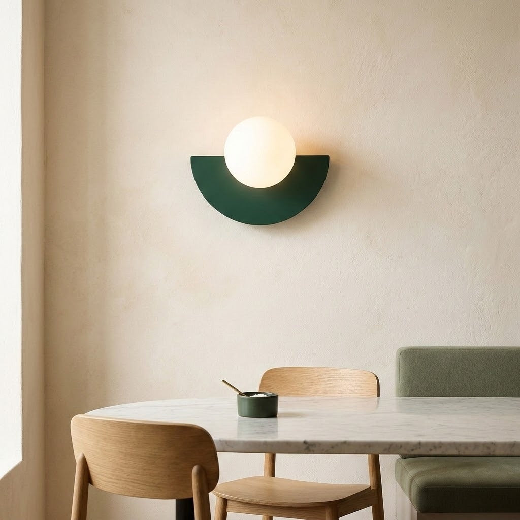 A modern semi-circular wall sconce with a dark green metal shade and a glowing white globe bulb, mounted on a textured beige wall above a dining table with wooden chairs.