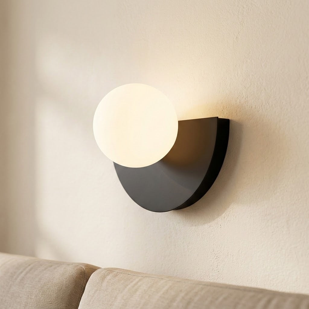 Black wall sconce with a glowing white globe, viewed obliquely to show its projection from a cream wall above a sofa.