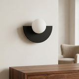 A modern semi-circular wall sconce with a matte black metal base and a white spherical glass shade, mounted on a light beige wall above a wooden console table.
