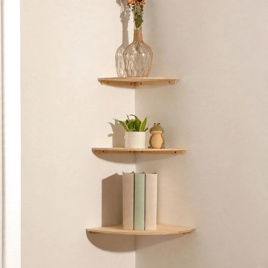 Three light natural pine corner shelves in a cream corner holding a flower vase, a potted plant and frog ornament, and three upright books.