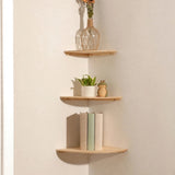 Three light natural pine corner shelves in a cream corner holding a flower vase, a potted plant and frog ornament, and three upright books.