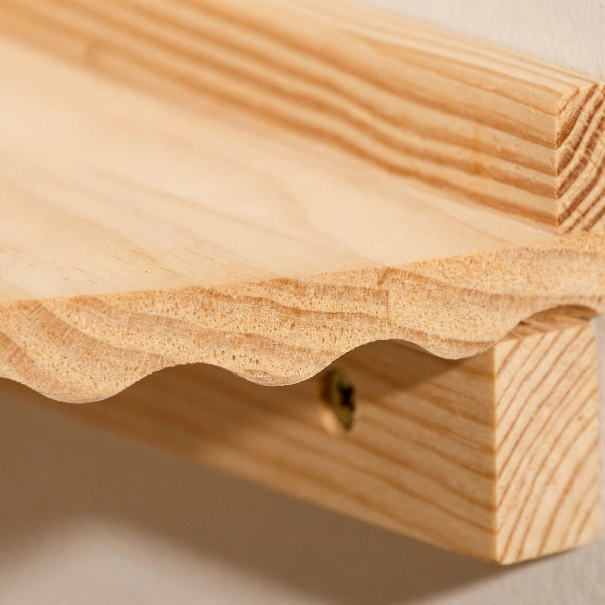 Extreme close-up of a light natural pine corner shelf showing its wavy scalloped front edge, wood grain, and wooden cleat with a screw below.