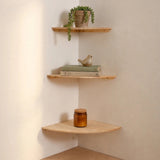 Three light natural wood corner shelves mounted on a cream-colored wall, styled with a trailing green plant, books and a small bird figurine, and an amber glass jar.