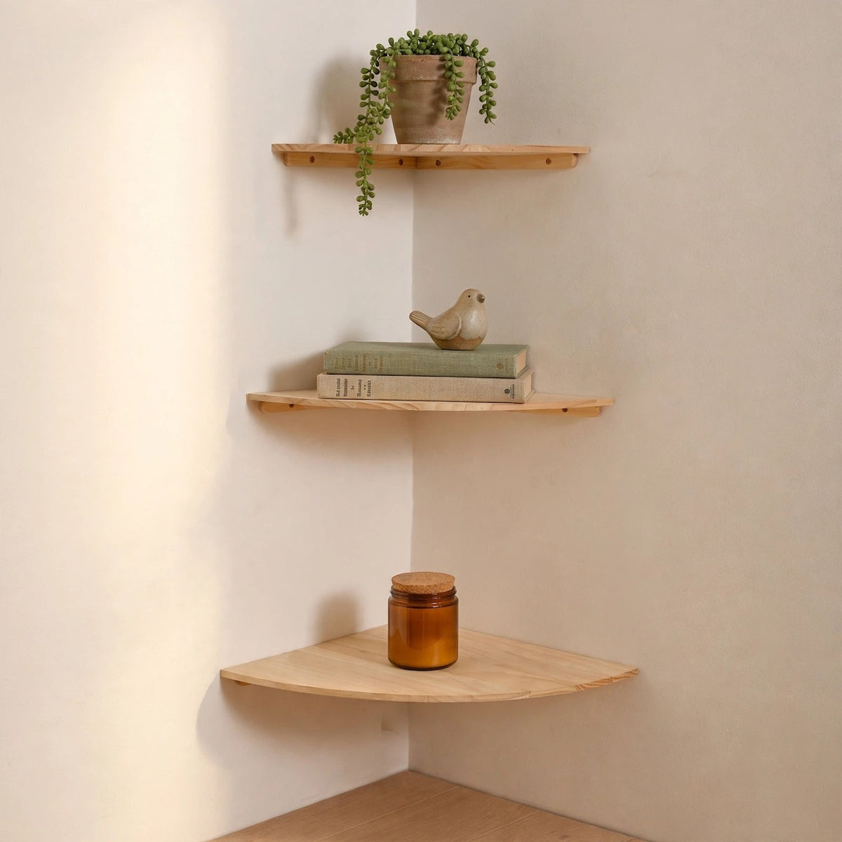 Three light natural wood corner shelves mounted on a cream-colored wall, styled with a trailing green plant, books and a small bird figurine, and an amber glass jar.