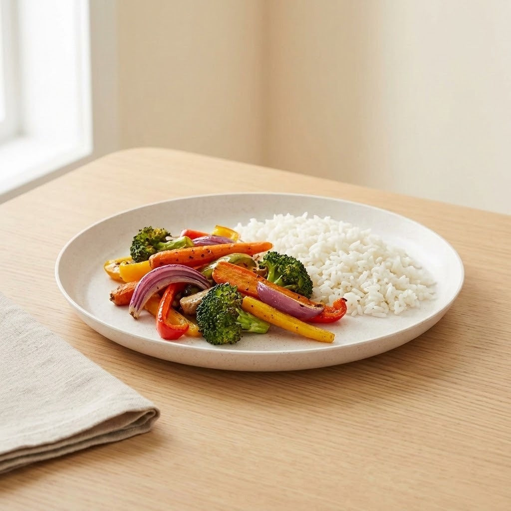 A round off-white ceramic plate with a subtle textured finish, containing white rice and cooked vegetables (broccoli, red onion, carrots, and bell peppers), set on a light wood table.