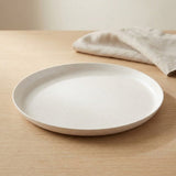 A round off-white ceramic plate with a textured matte finish, set on a light wood table with a beige linen napkin in the background.