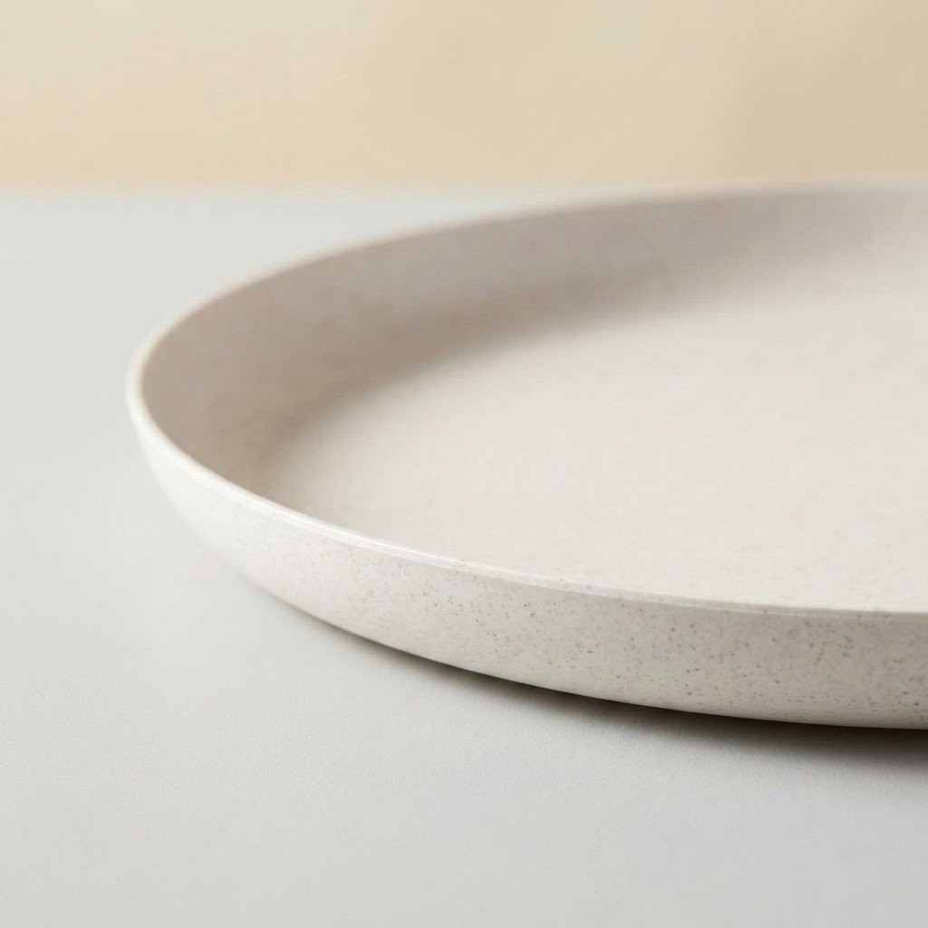 A close-up of a shallow round plate with a speckled off-white finish, resting on a light surface against a soft beige background.