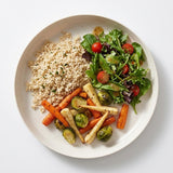 A top-down view of a light beige ceramic plate containing a salad with mixed greens, cherry tomatoes, roasted carrots, parsnips, and Brussels sprouts, alongside cooked brown rice on a white background.