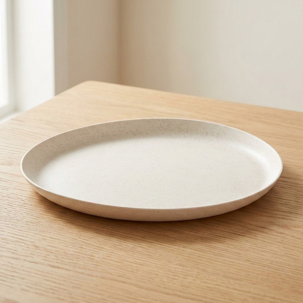 A round, shallow ceramic plate with a speckled cream finish, shown on a light wood table in a softly lit indoor setting.