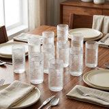 A set of clear textured glass tumblers arranged on a wooden dining table with plates, napkins, and silverware in a real dining room setting.