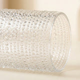 A close-up of a clear textured glass cylinder with a raised bubble or dimple pattern, photographed on a soft warm beige background.