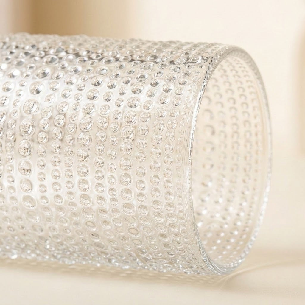 A close-up of a clear textured glass cylinder with a raised bubble or dimple pattern, photographed on a soft warm beige background.