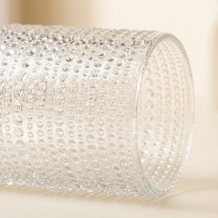 A close-up of a clear textured glass cylinder with a raised bubble or dimple pattern, photographed on a soft warm beige background.