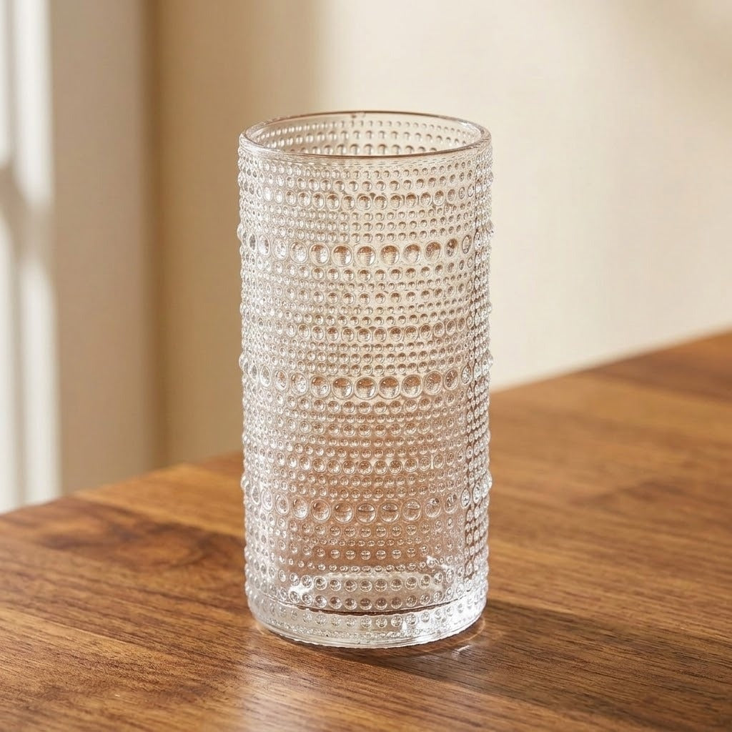 A clear textured glass tumbler with horizontal rows of raised dots, standing on a wooden tabletop against a softly blurred beige background.