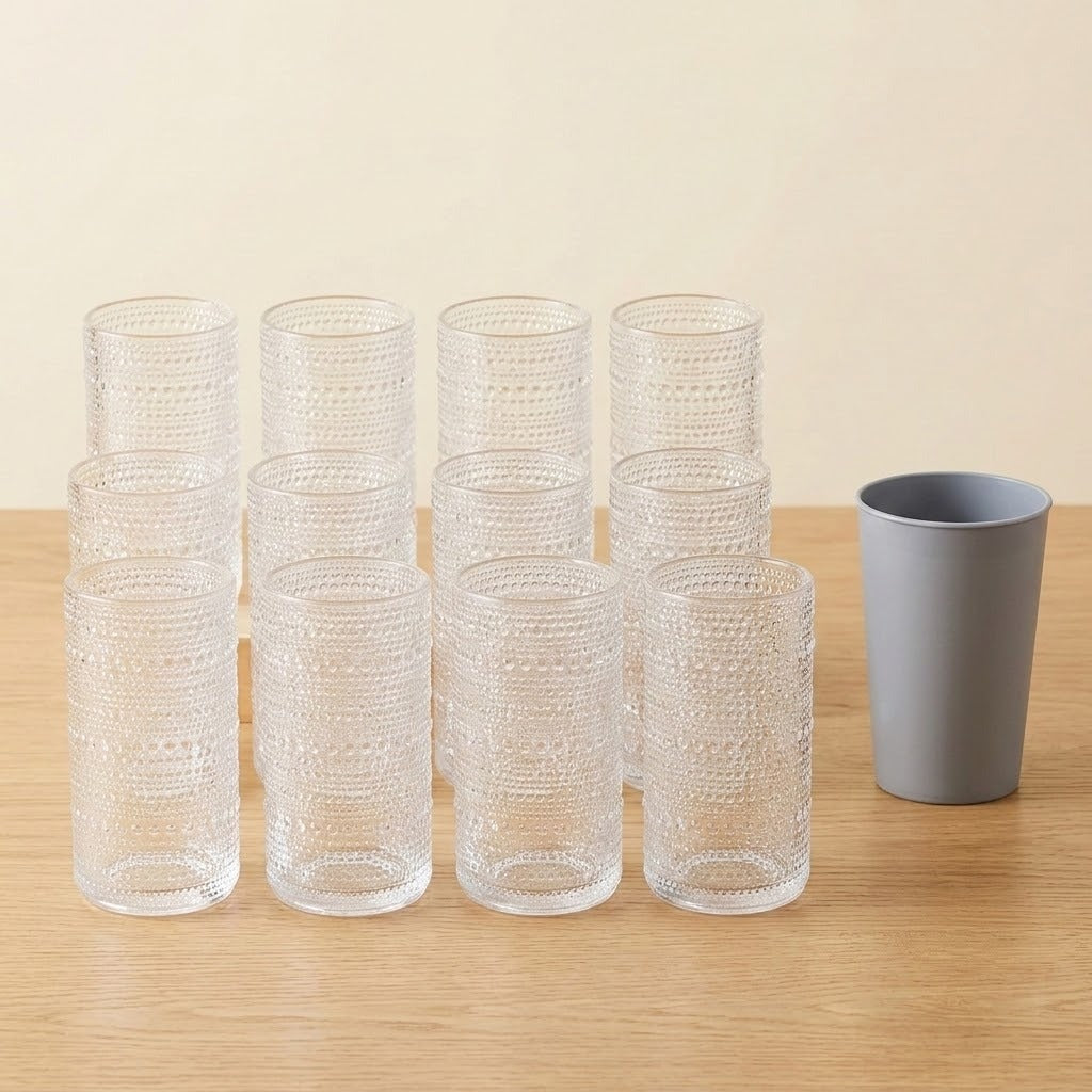 Nine clear textured drinking glasses stacked in two rows on a light wood table, with a single gray cup standing to the right, against a warm beige background.