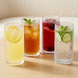 Four textured clear drinking glasses filled with iced beverages on a light wood surface, with a soft neutral indoor background.