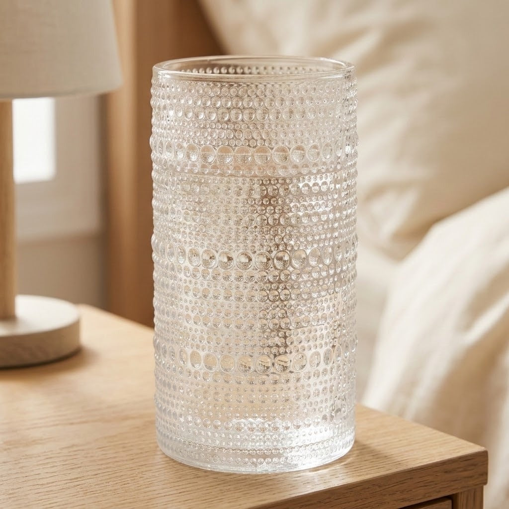 A clear textured glass vase with a beaded bubble pattern, sitting on a light wood bedside table in a softly lit bedroom setting.