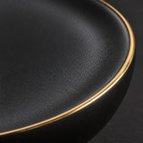 Macro detail of the matte black ceramic texture and narrow gold rim on the dinnerware.