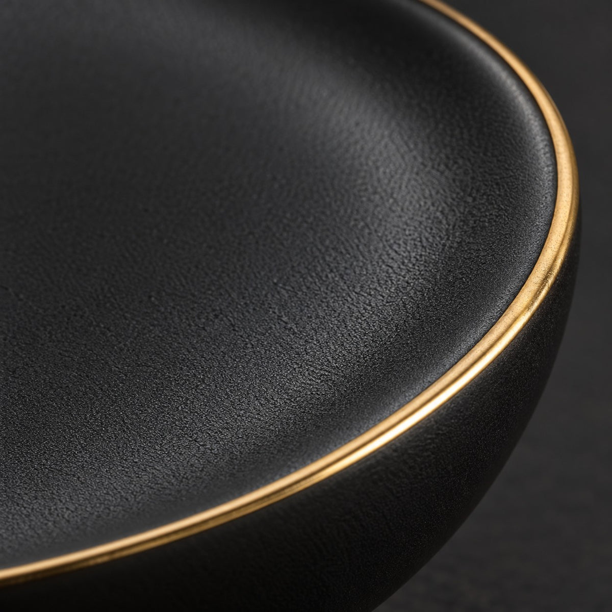 Macro detail of the matte black ceramic texture and narrow gold rim on the dinnerware.
