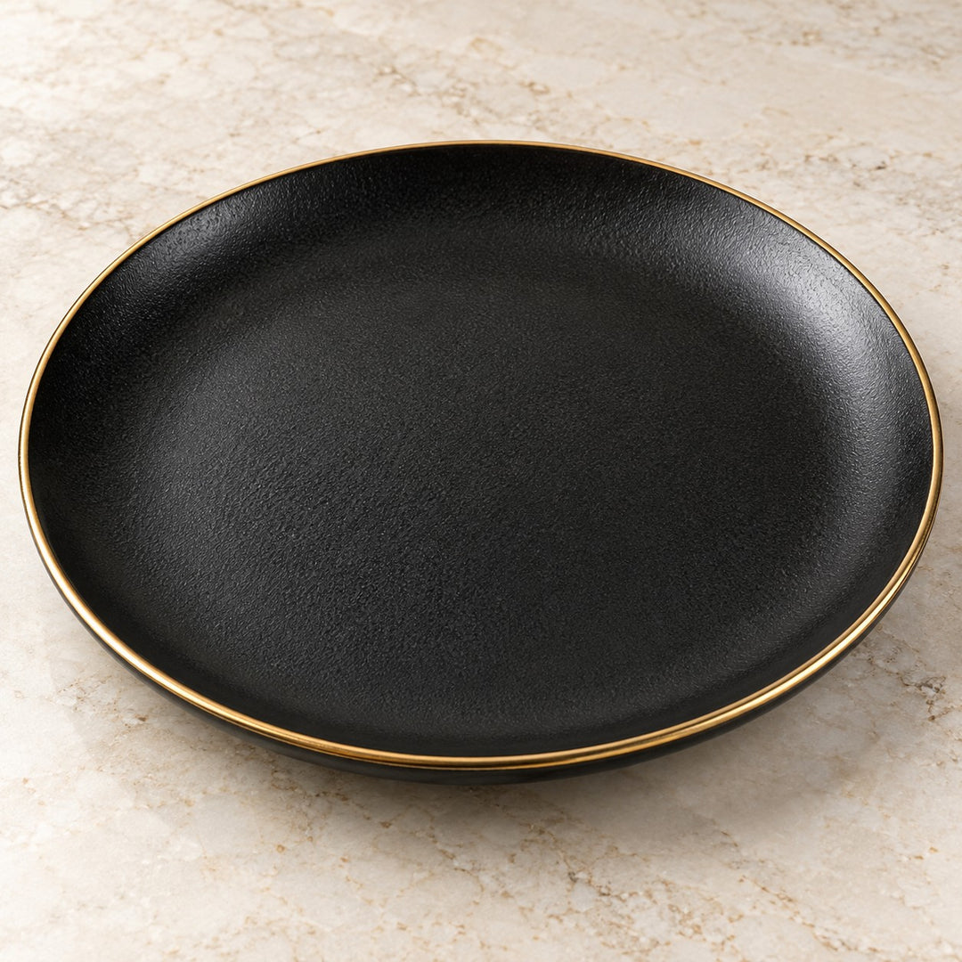 Matte black ceramic dinner plate with a fine gold rim shown large on a pale kitchen surface.