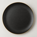 Single shallow black ceramic plate with a delicate gold rim centered on a light studio background.